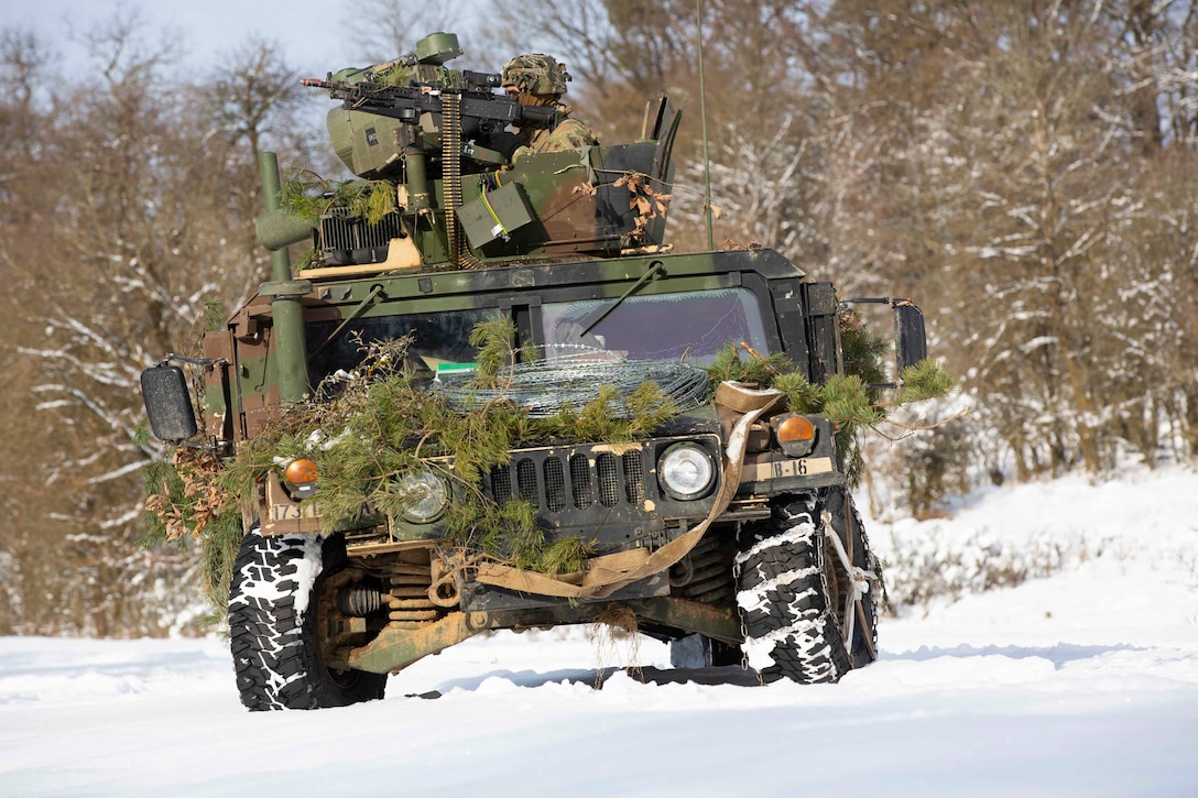 Soldiers ride in a Humvee in the snow; trees can be seen in the background.