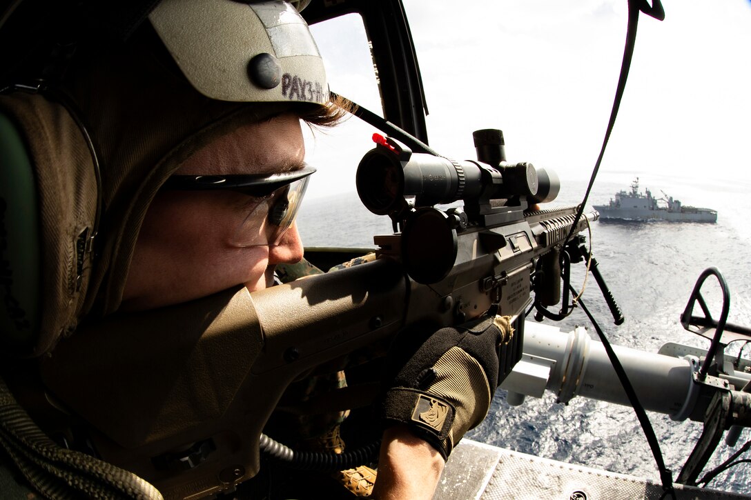 A Marine aims a weapon at a ship from a helicopter.