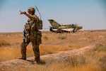 A soldier stands in a dry field and points at something in the distance; behind him, an old aircraft sits rusting in the desert.