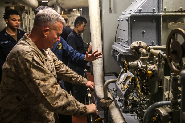 A man in a military uniform looks at equipment in a small, enclosed space; four men in uniforms are in the background.