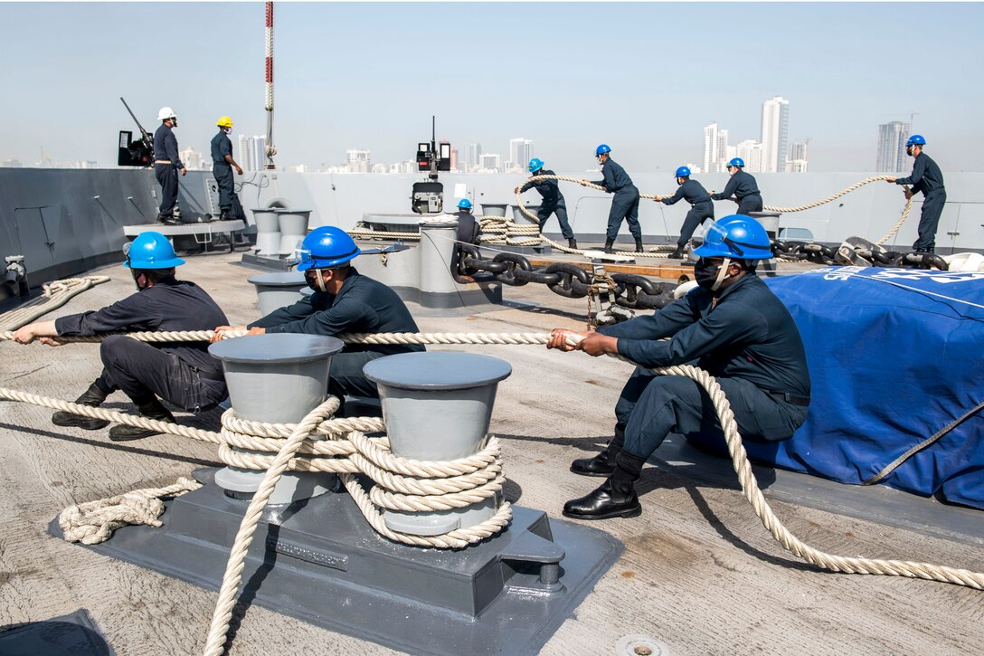 Sailors in a line on a ship pull a rope, while in the background another group pulls a different rope.