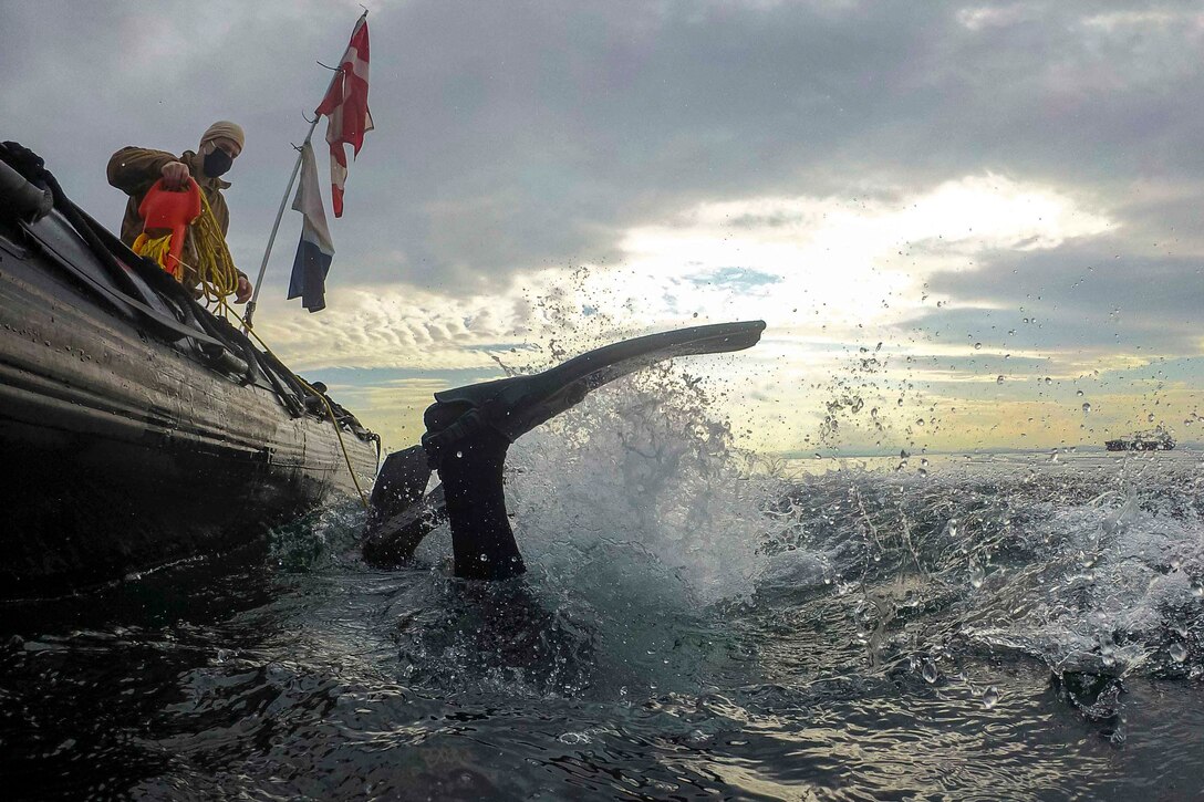 A man stands inside a small inflatable boat as a sailor dives into a water.