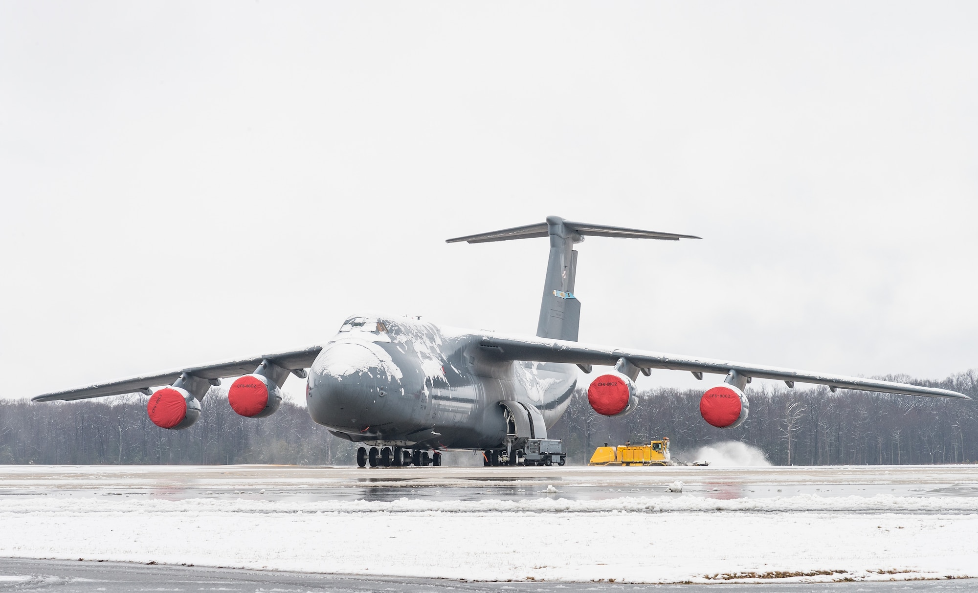 A snow-covered C-5M Super Galaxy sits on the flight line at Dover Air Force Base, Delaware, Feb. 7, 2021. As snow fell, the base continued normal operations and prepared for additional snowfall. In the background, 436th Civil Engineer Squadron snow removal teams plowed snow off the airfield and flight line. (U.S. Air Force photo by Roland Balik)