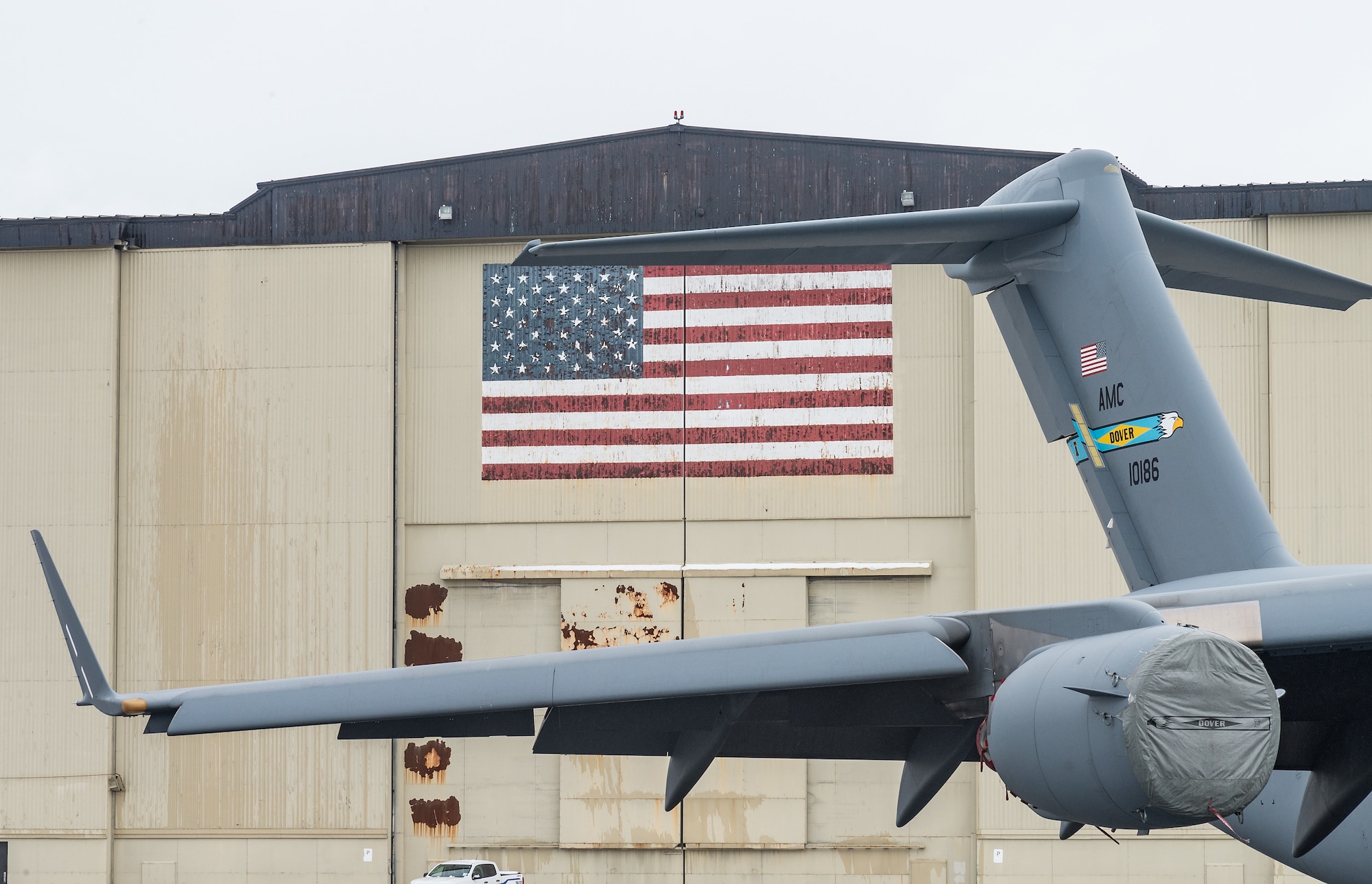 While in tow, a C-17 Globemaster III t-tail passes by the U.S. flag painted on a hangar at Dover Air Force Base, Delaware, Feb. 7, 2021. The aircraft was towed into a hangar to facilitate maintenance out from the weather. (U.S. Air Force photo by Roland Balik)