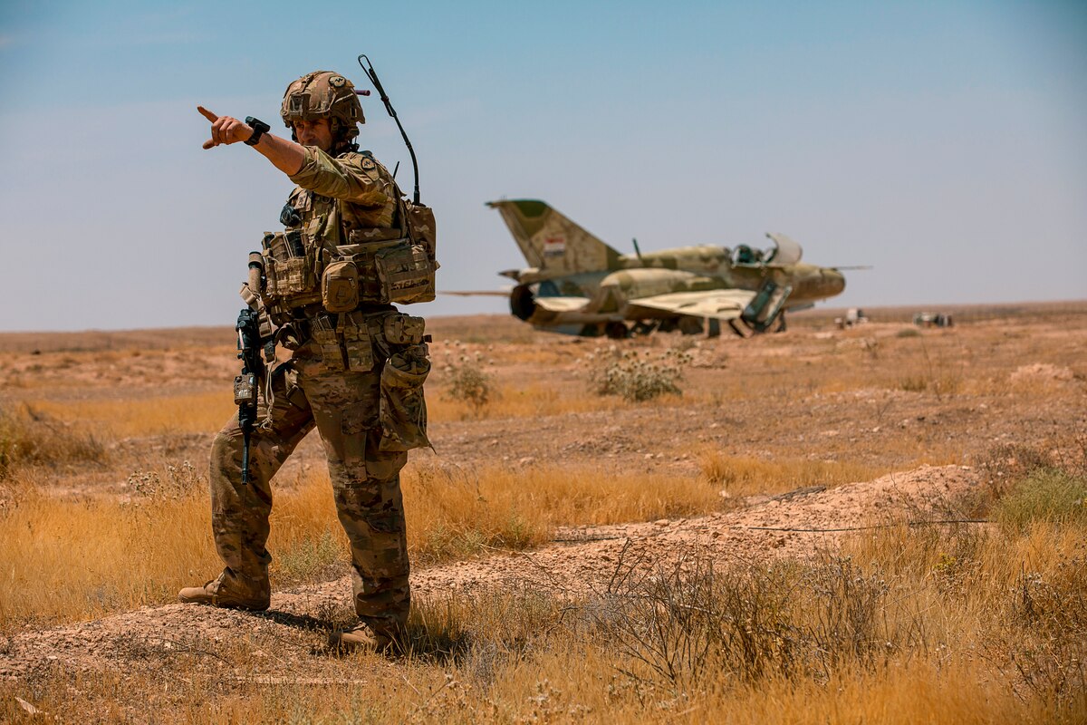 A soldier stands in a dry field and points at something in the distance; behind him, an old aircraft sits rusting in the desert.