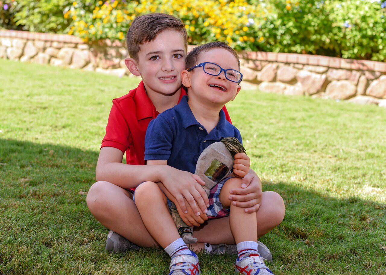 A toddler sits in his brother's lap on the grass as the two pose for a photo.