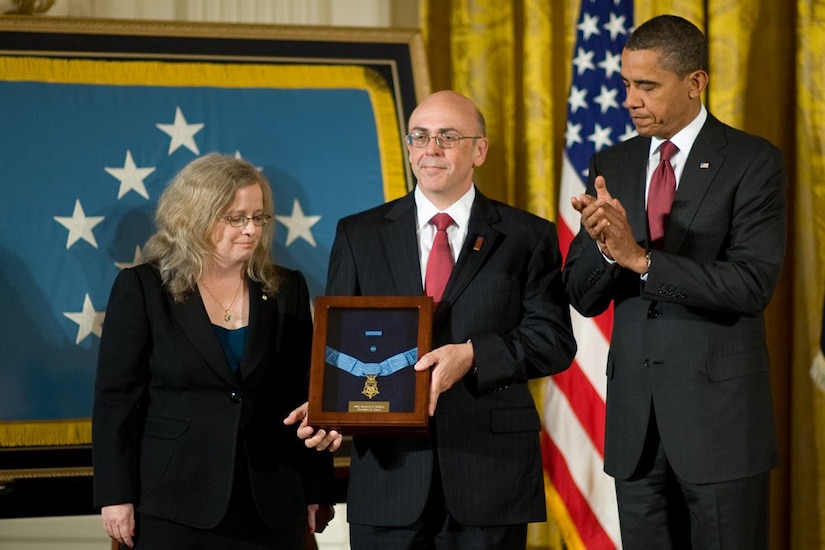 A man holds a medal in a box. On the left, a woman looks down. On the right, a man applauds.