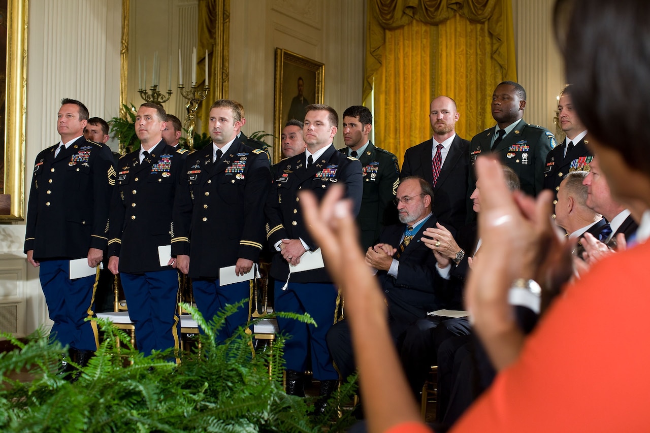 A woman in the foreground applauds as soldiers stand at attention in the background. Others in the audience also applaud.