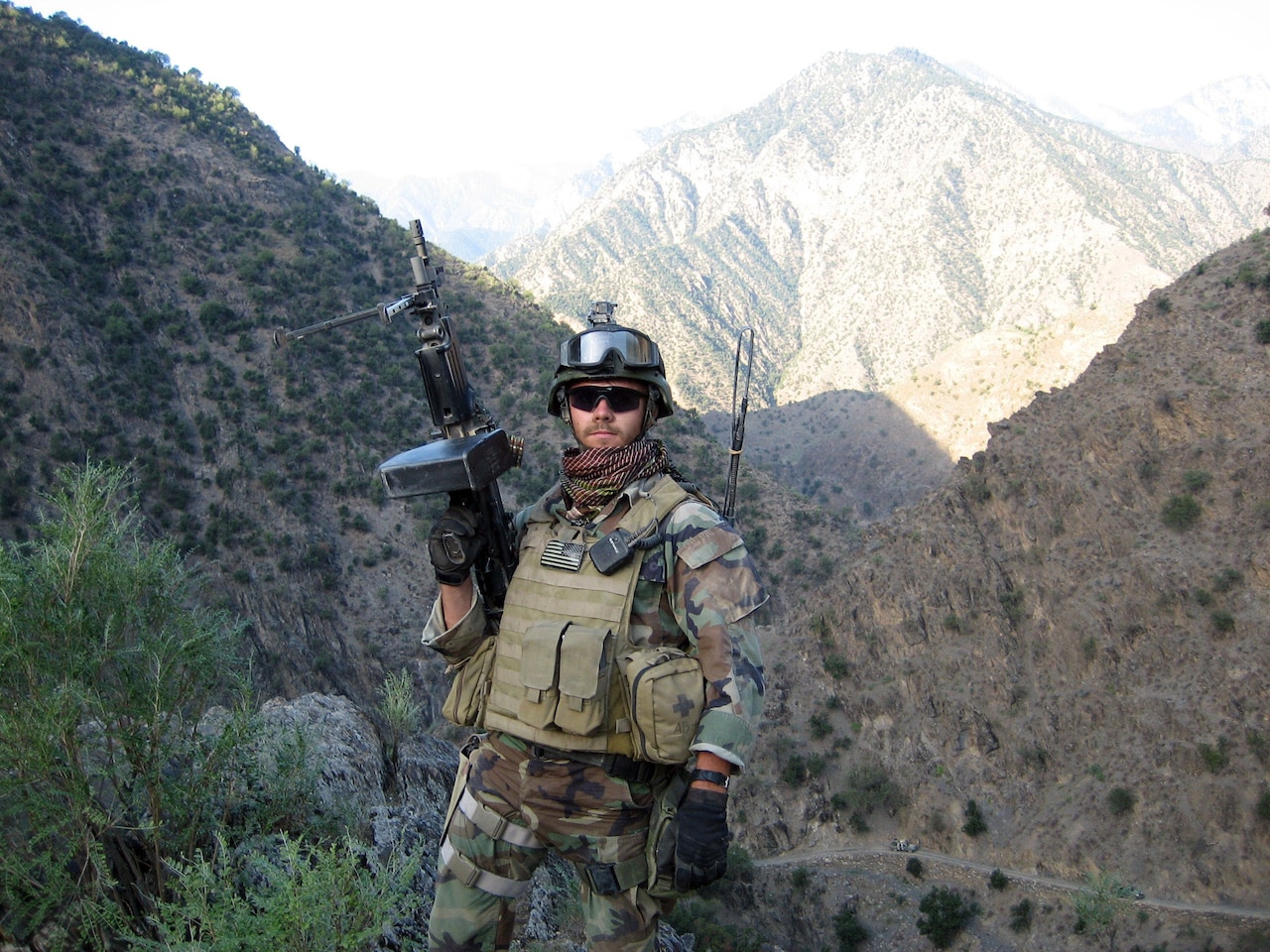 A man in combat gear points a large weapon into the air. Mountains line the background.