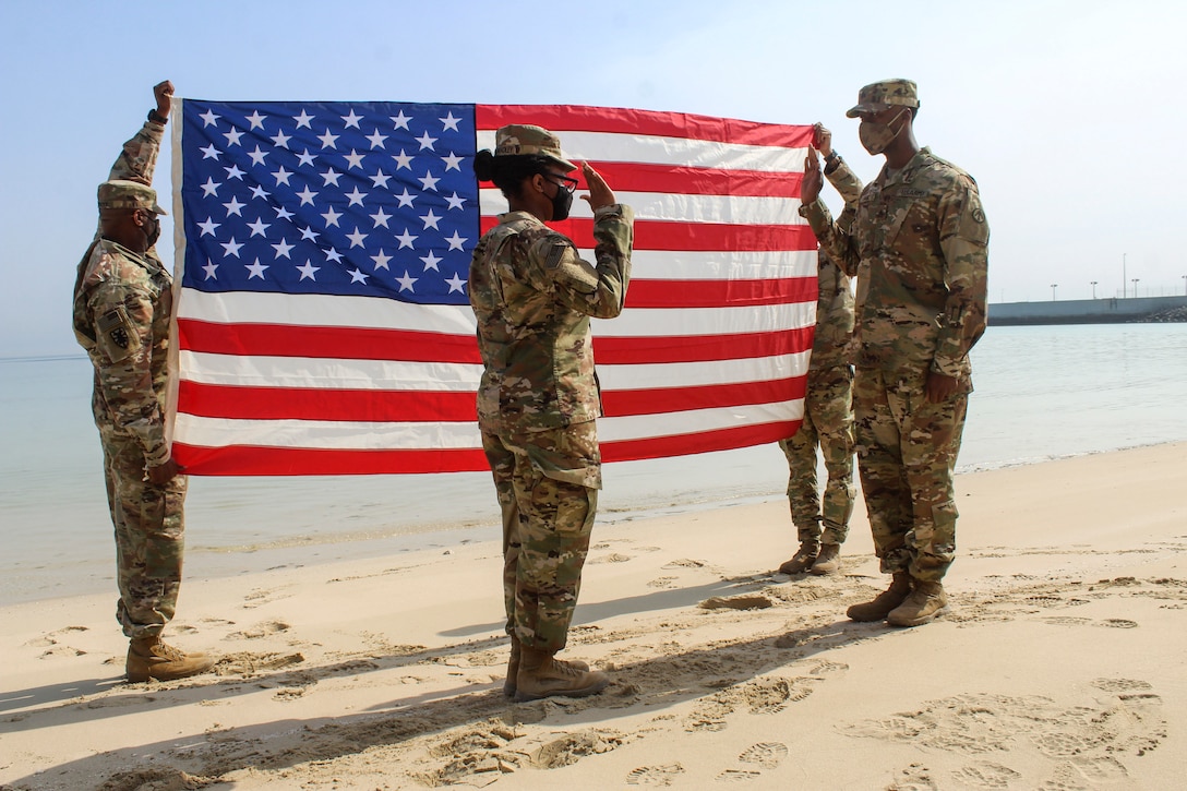 Two soldiers face each other with right hands raised on a beach, as two others hold up an American flag behind them.