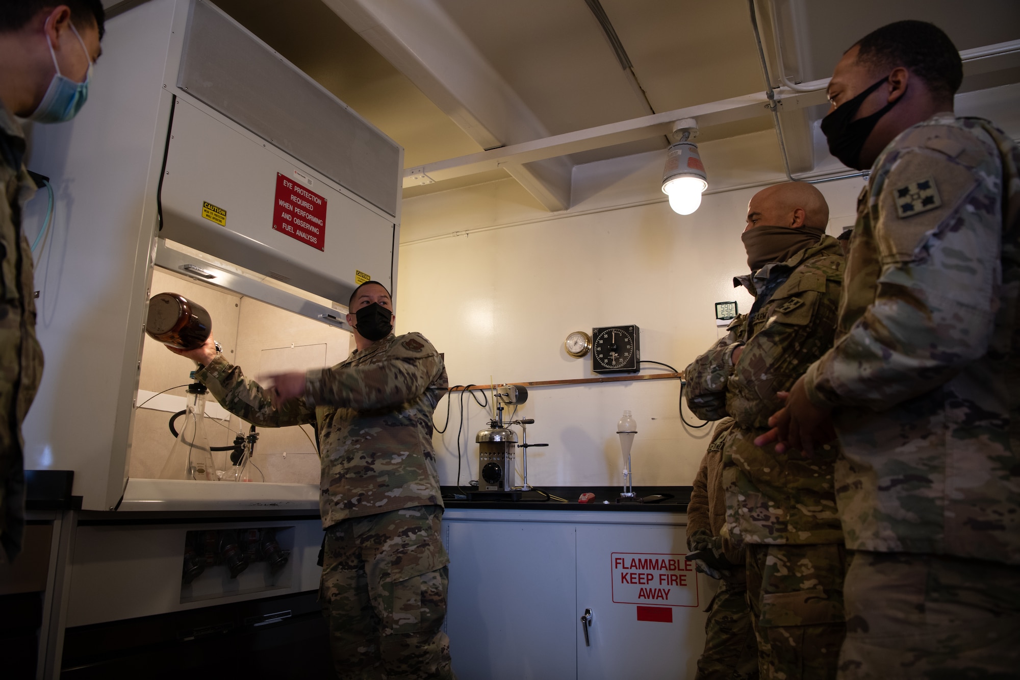 An Airman holds a brown jar inside of a specialized aircraft fuel testing machine, speaking in a small room to two soldiers.