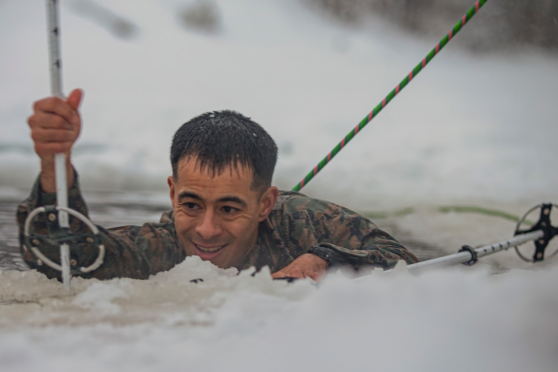 A Marine uses tools to break ice while immersed in an icy body of water.