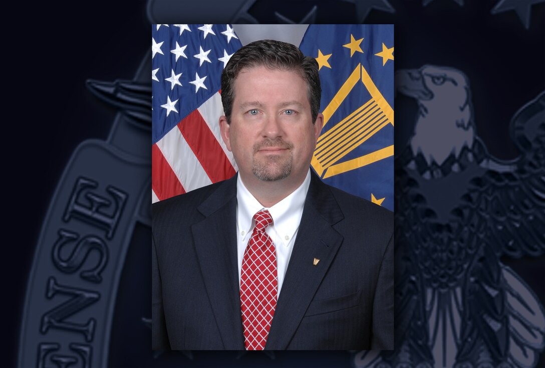 White man in a black suit, white shirt and red/white tie sits in front of the US and DLA flags.