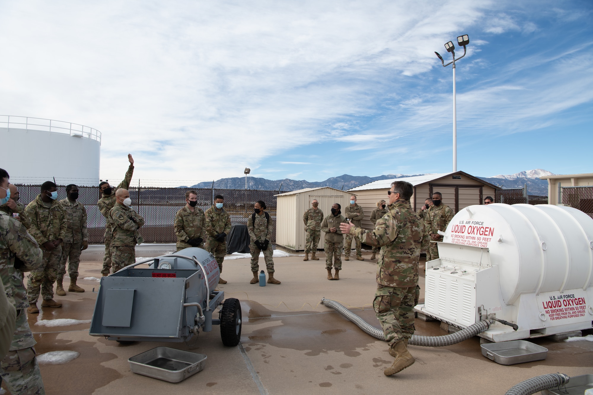 An Airmen speaks to a small group of soldiers in front of a large white container labeled "LIQUID OXYGEN" and a gray container beside him.