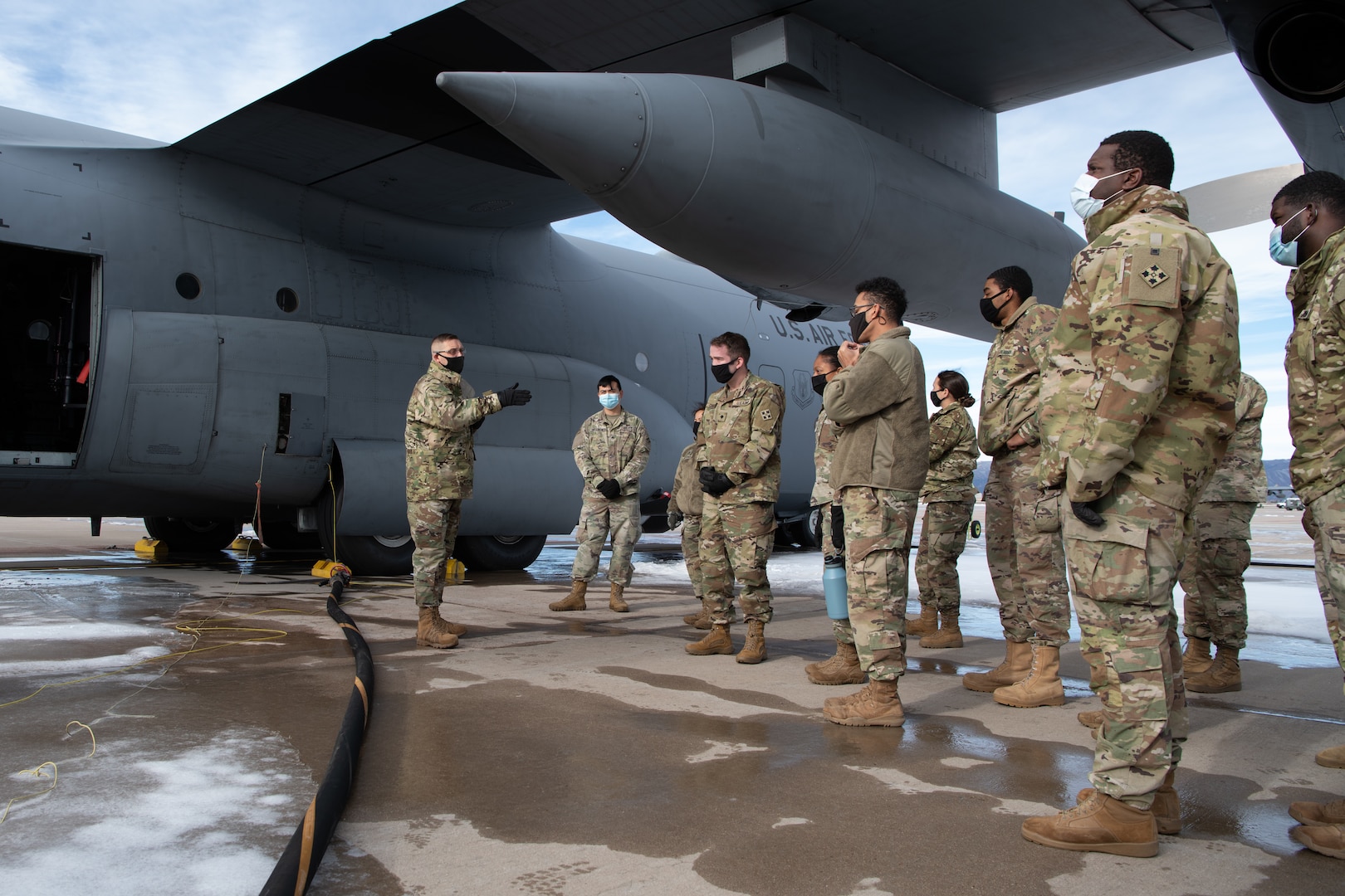 A man speaks to a small group of soldiers in front of a C-130 aircraft with a black fuel hose on the ground leading to the aircraft.