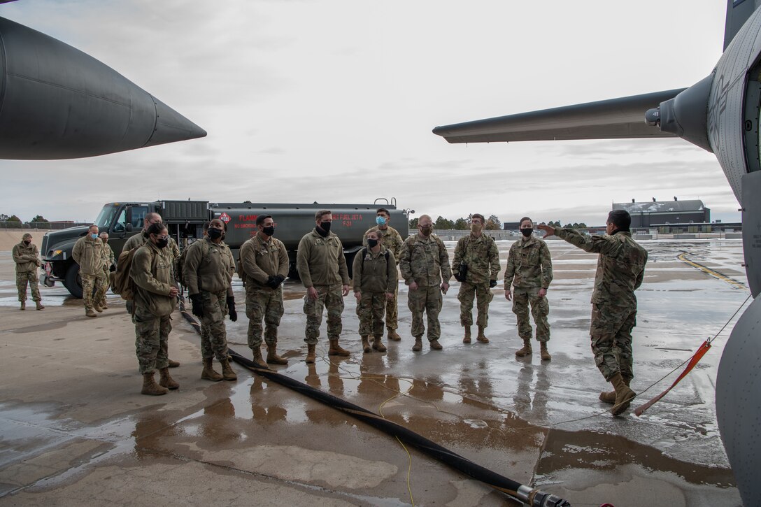 An Airman speaks to a small group of soldiers on the flight line with a fuel truck in the background and a C-130 in the foreground with a large fuel hose leading from the truck to aircraft.