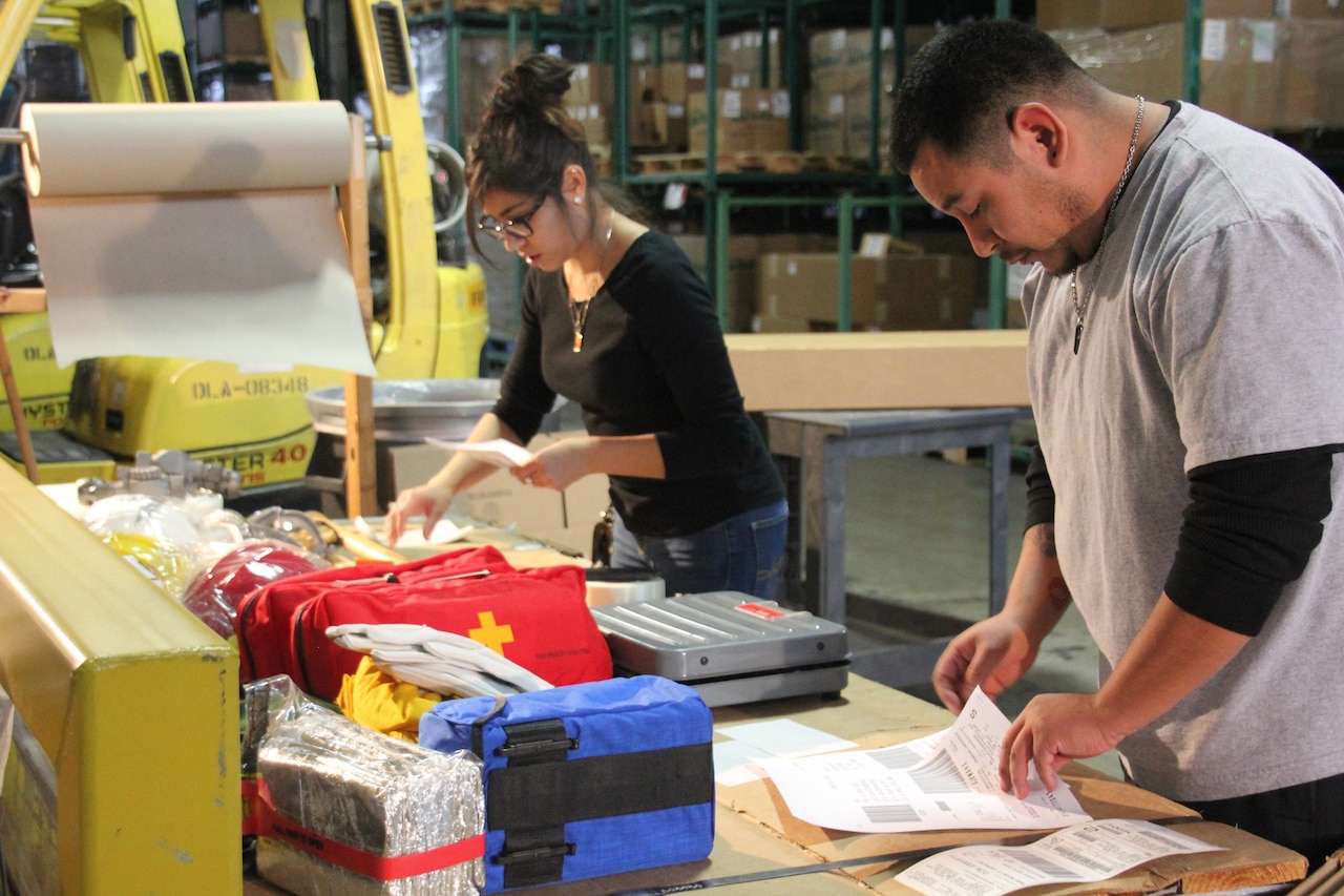 A woman and a man read documents as they stand at a table in a crowded warehouse.