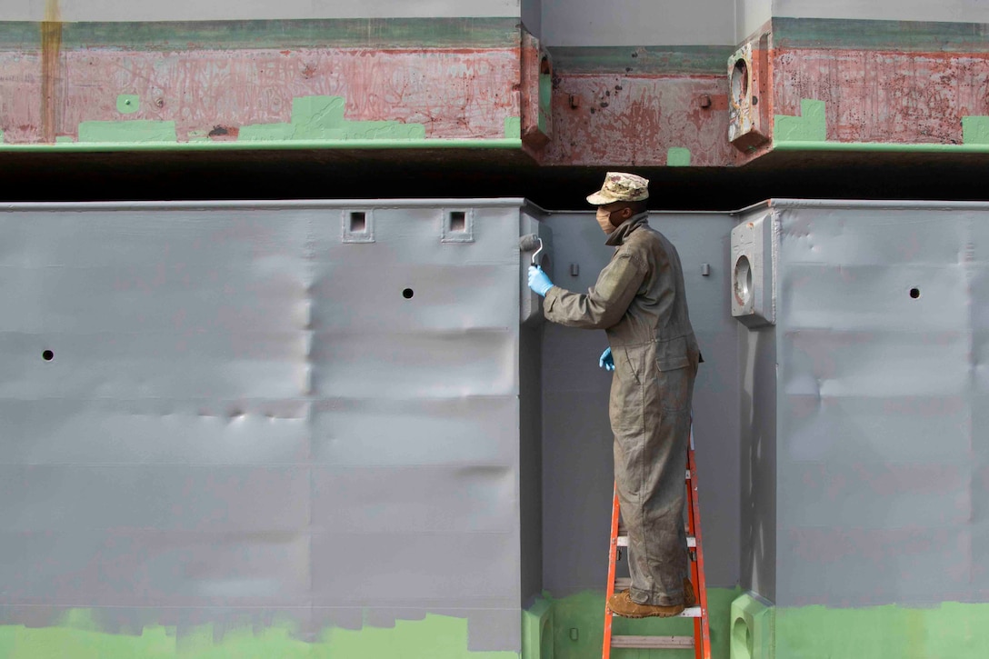 A sailor stands on a ladder while painting the hull of a vessel.