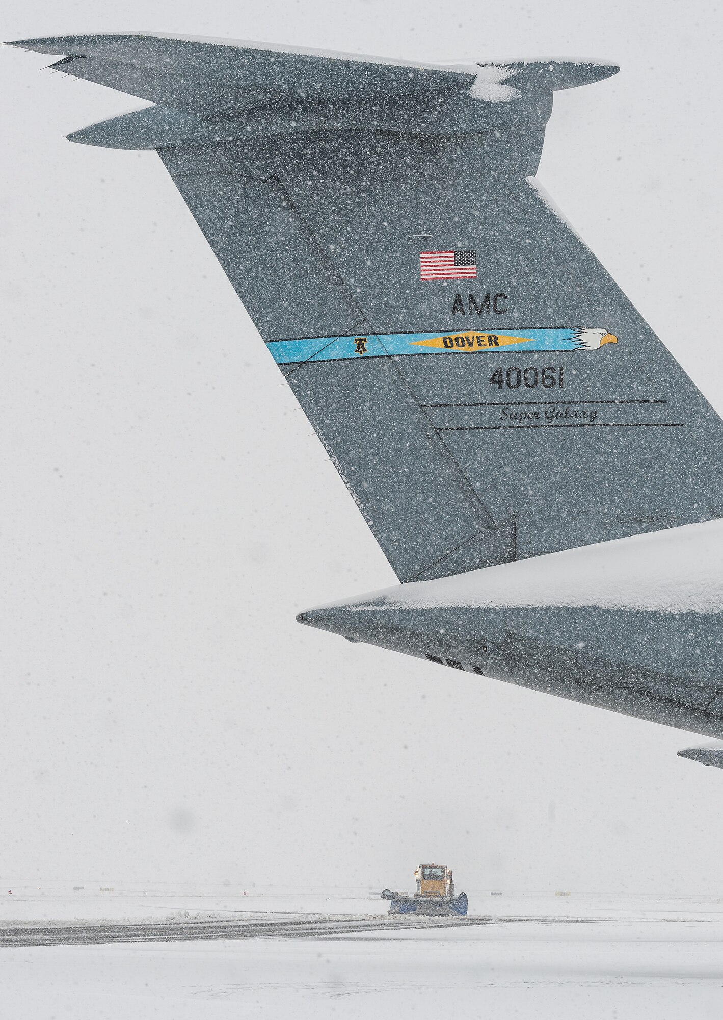 A snow-covered C-5M Super Galaxy tail appears to hang over a 436th Civil Engineer Squadron snowplow as it clears a taxiway at Dover Air Force Base, Delaware, Feb. 11, 2021. As Winter Storm Roland produced a wintry mix of precipitation, the base continued normal operations and prepared for additional forecast snowfall. (U.S. Air Force photo by Roland Balik)