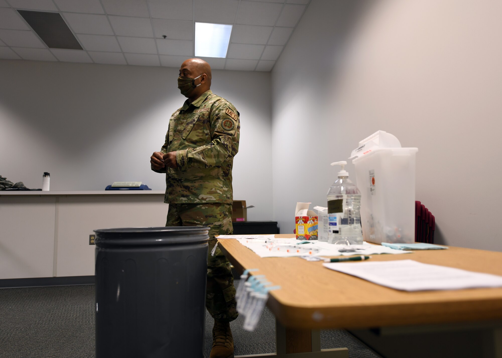A US Air Force Master Sgt. stands next to a table with vaccine materials, including needles and vials of medication.