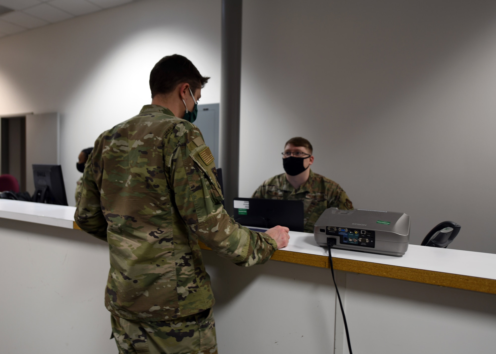A masked Airman stands at a counter and another masked Airman sits behind it.