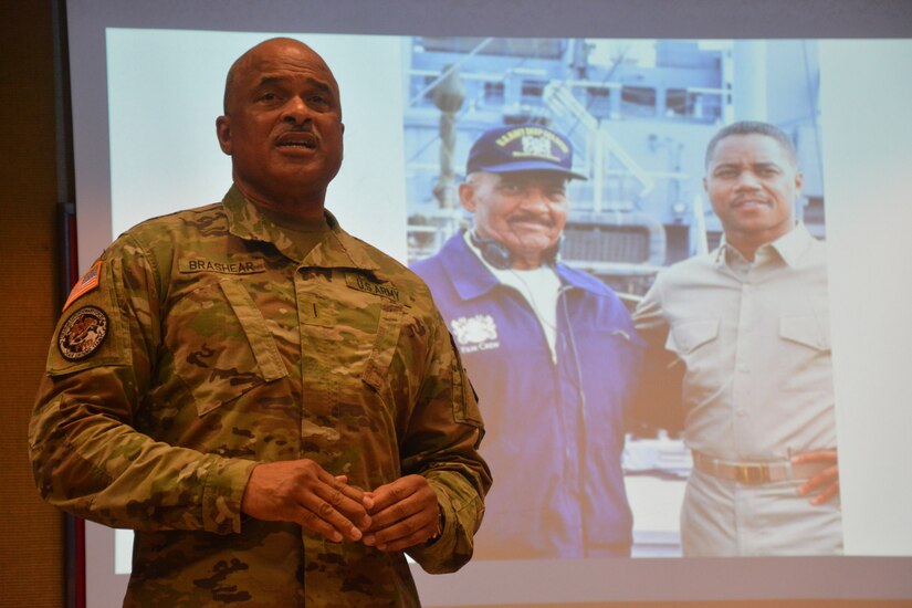 A soldier stands and speaks in front of a photo of his father projected on a screen.