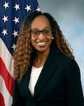 Sabrina in a black suit jacket with a white shirt in front of the flag and blue background.