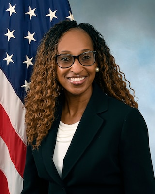 Sabrina in a black suit jacket with a white shirt in front of the flag and blue background.