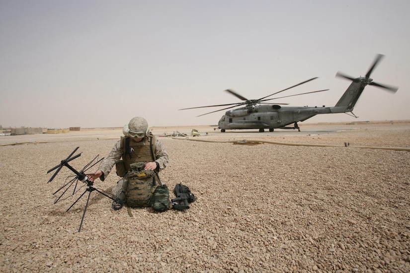A service member keels on a bed of rocks to work on a satellite communications device; a helicopter is in the background.