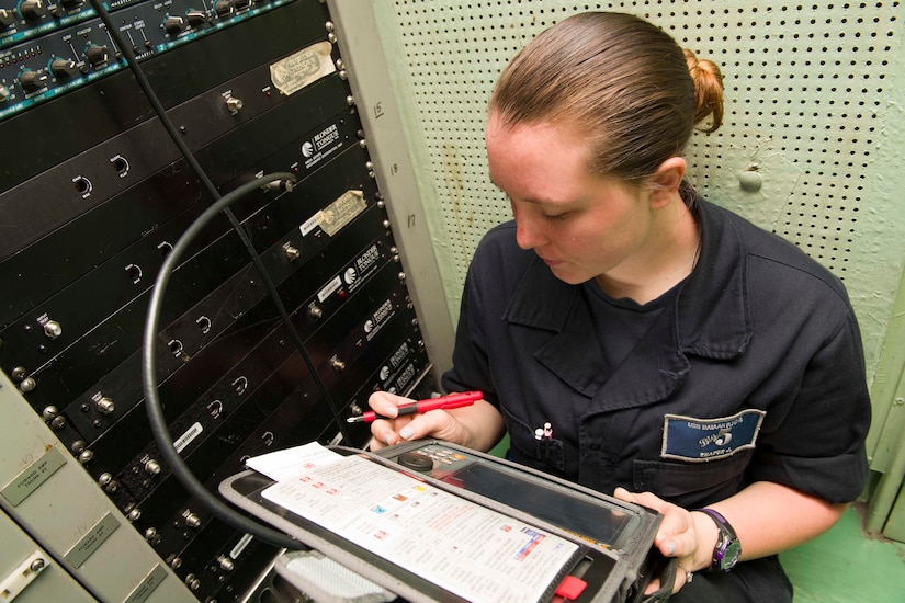 A woman dressed in a military uniform sits as she looks at a small on screen that is hooked up to a large panel to her right.