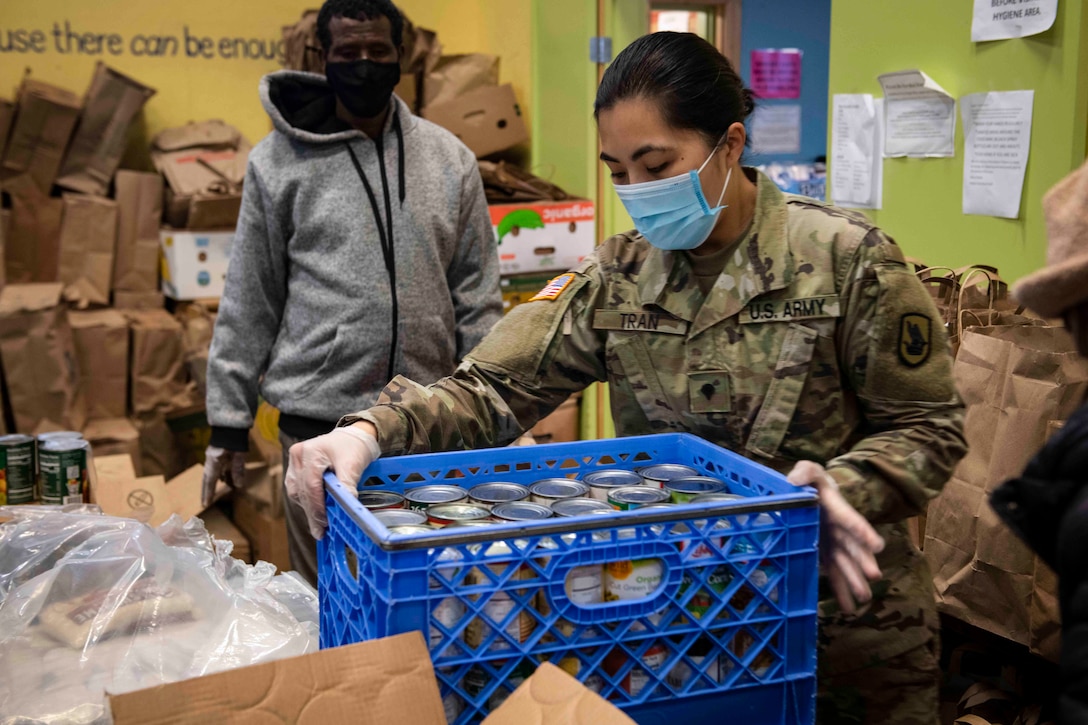 A soldier holds onto a crate of canned as another person stands behind.