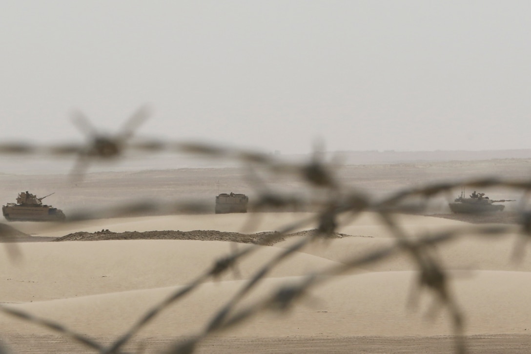 Tanks drive on the sand as seen through barbed wire.
