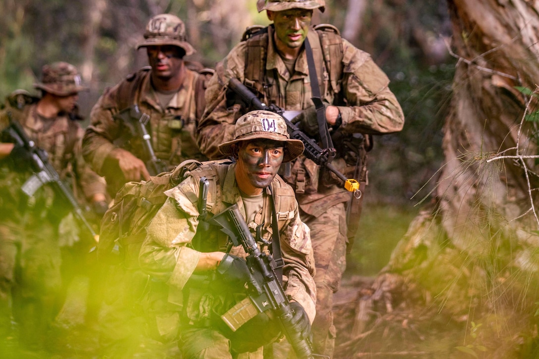 Soldiers dressed in camouflage move through the woods while carrying weapons.