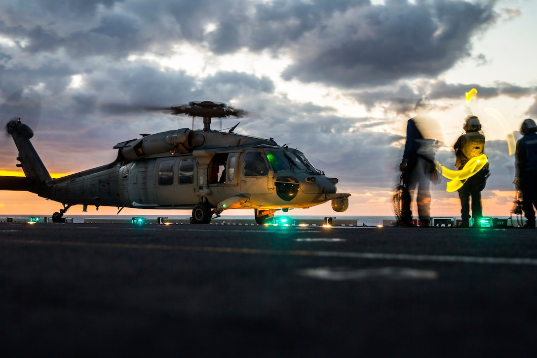 Marines and sailors stand near a helicopter on a ship illuminated by blue, green and yellow lights.