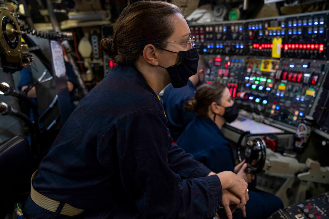 A sailor stands inside the control room of a ship; others sit behind.