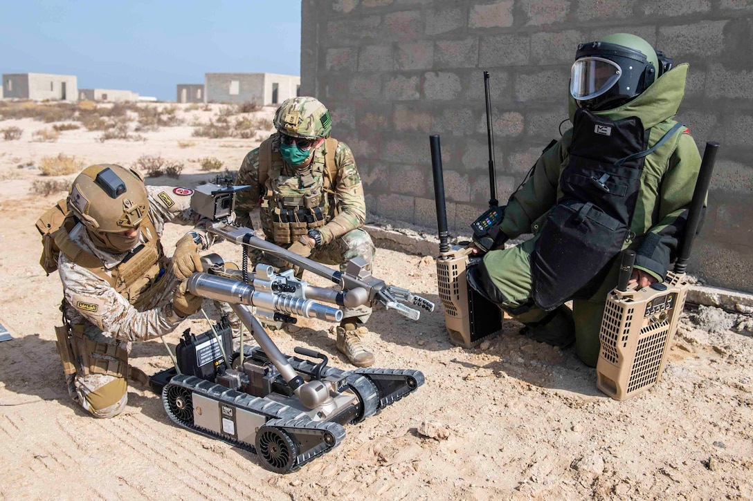 A sailor kneels on the ground watching a service member work on a robot; another person kneels beside wearing bomb protection gear.
