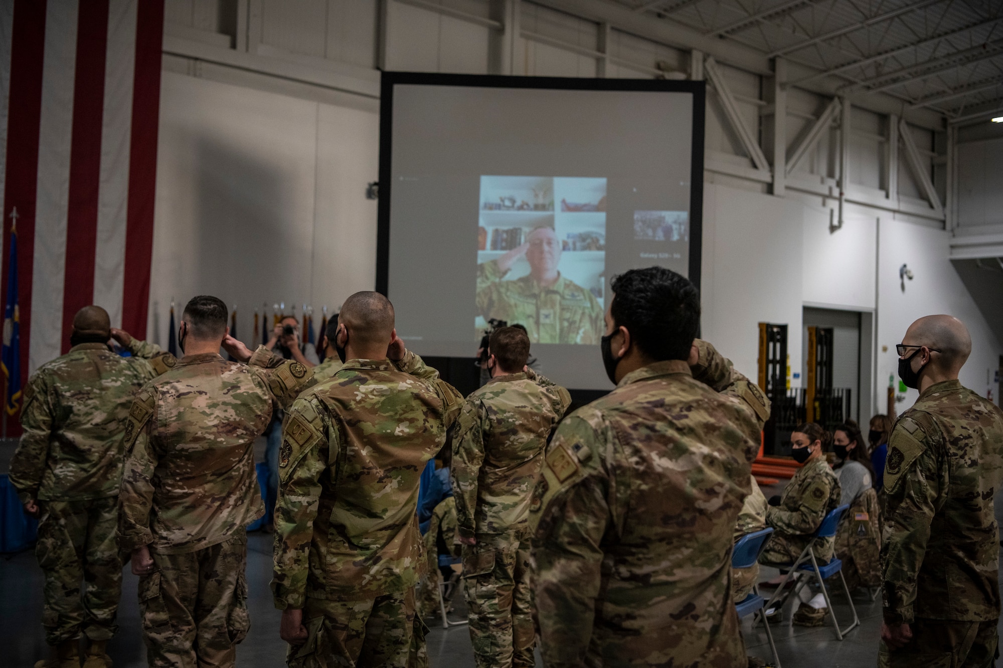 Men and Women salute a Colonel that is broadcasting in from a zoom call.