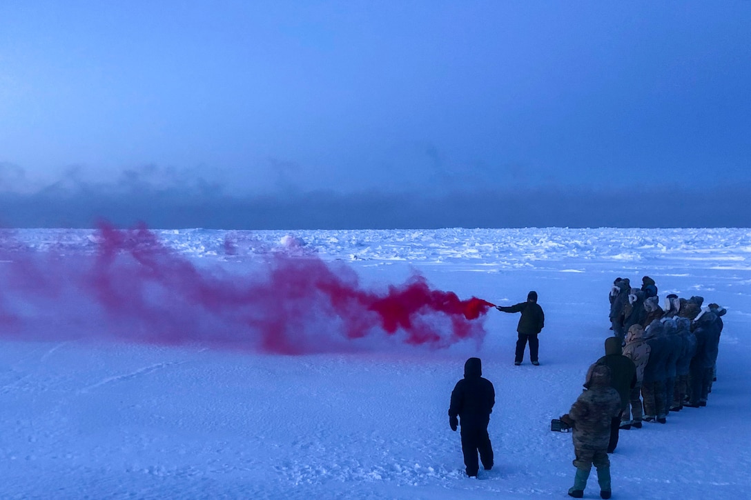 Service members watch as a streak of red smoke appears from an airman using a flare.