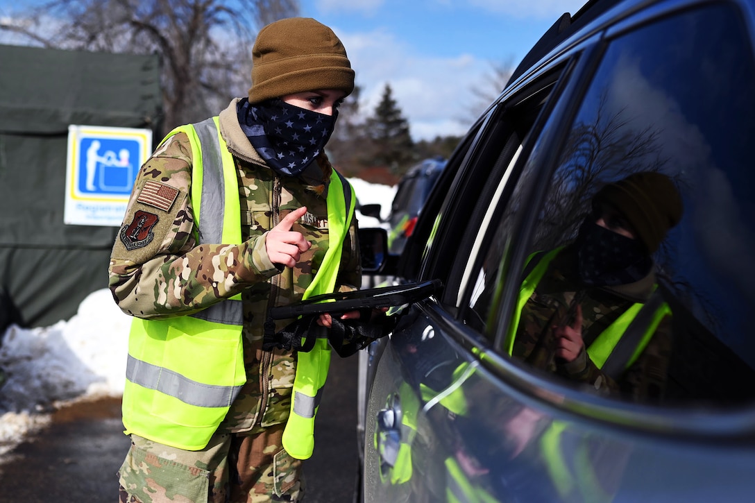An airman wearing a face mask holds a tablet and talks to someone in a vehicle.