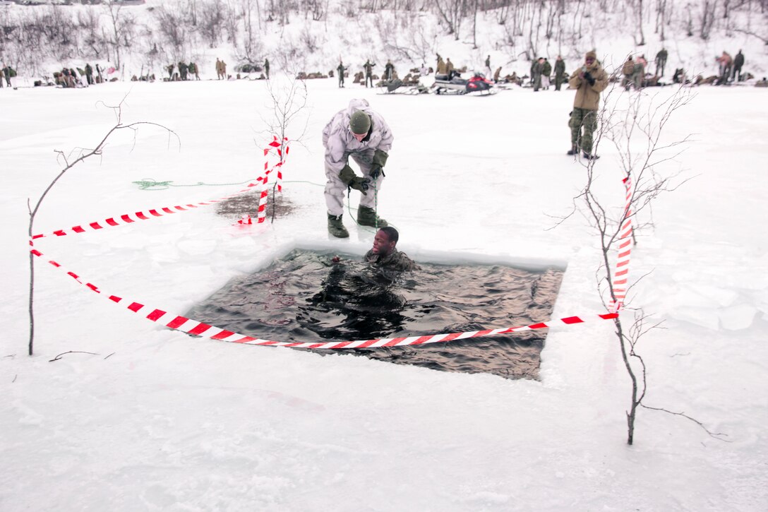 A service member looks at a watch while a Marine sits in a freezing water surrounded by snow.