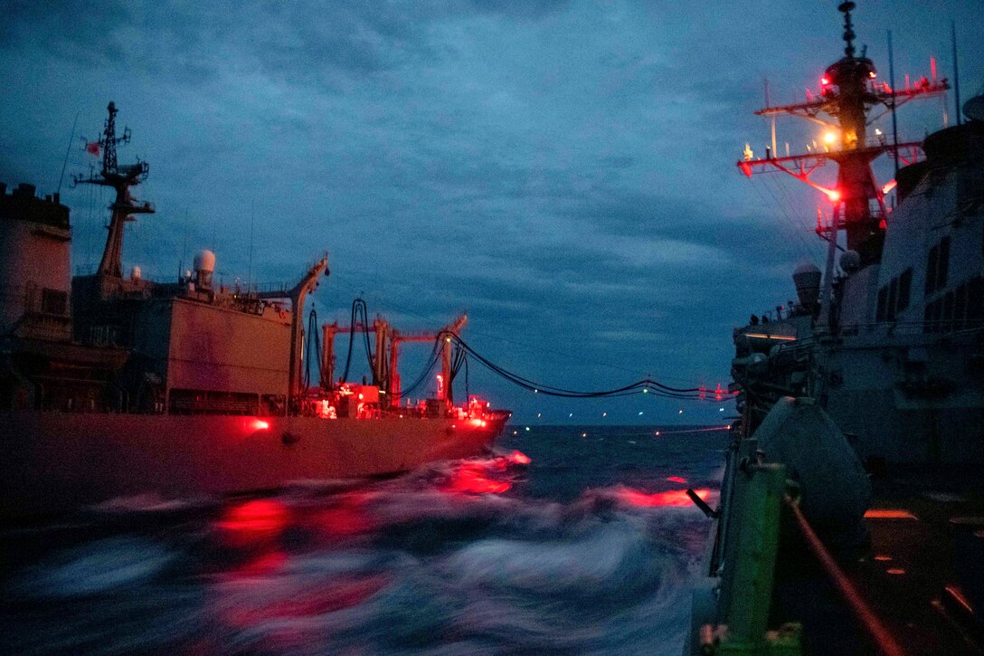 Two ships conduct a replenishment at sea illuminated by red lights.