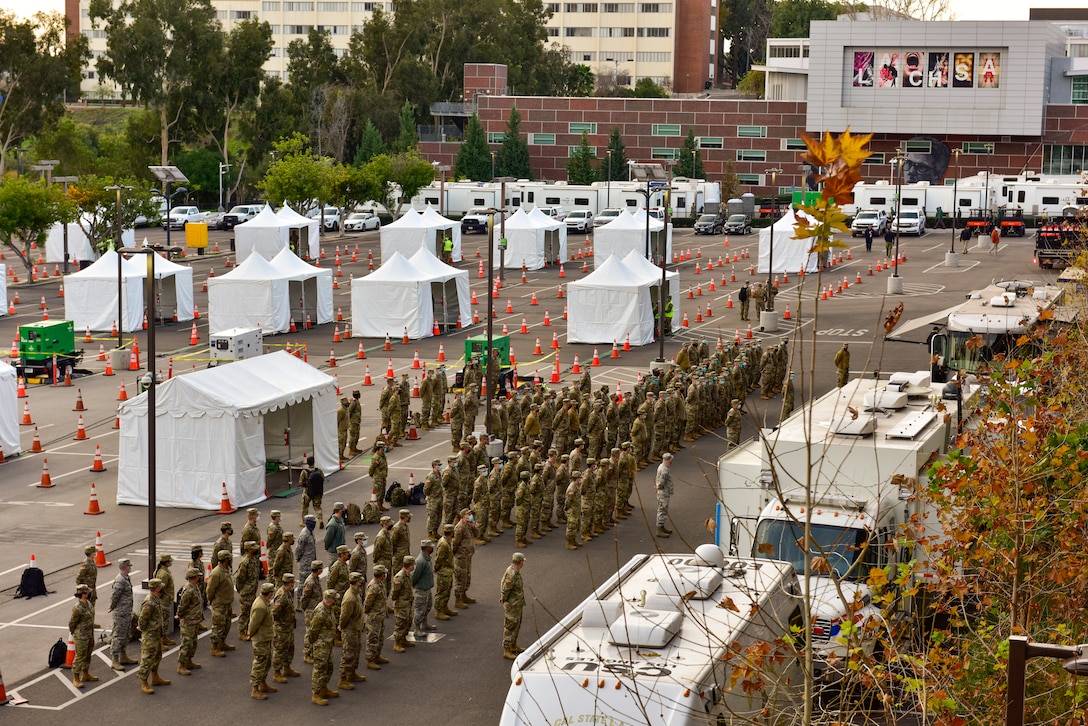 Dozens of service members stand in formation in a parking lot near an array of white tents.