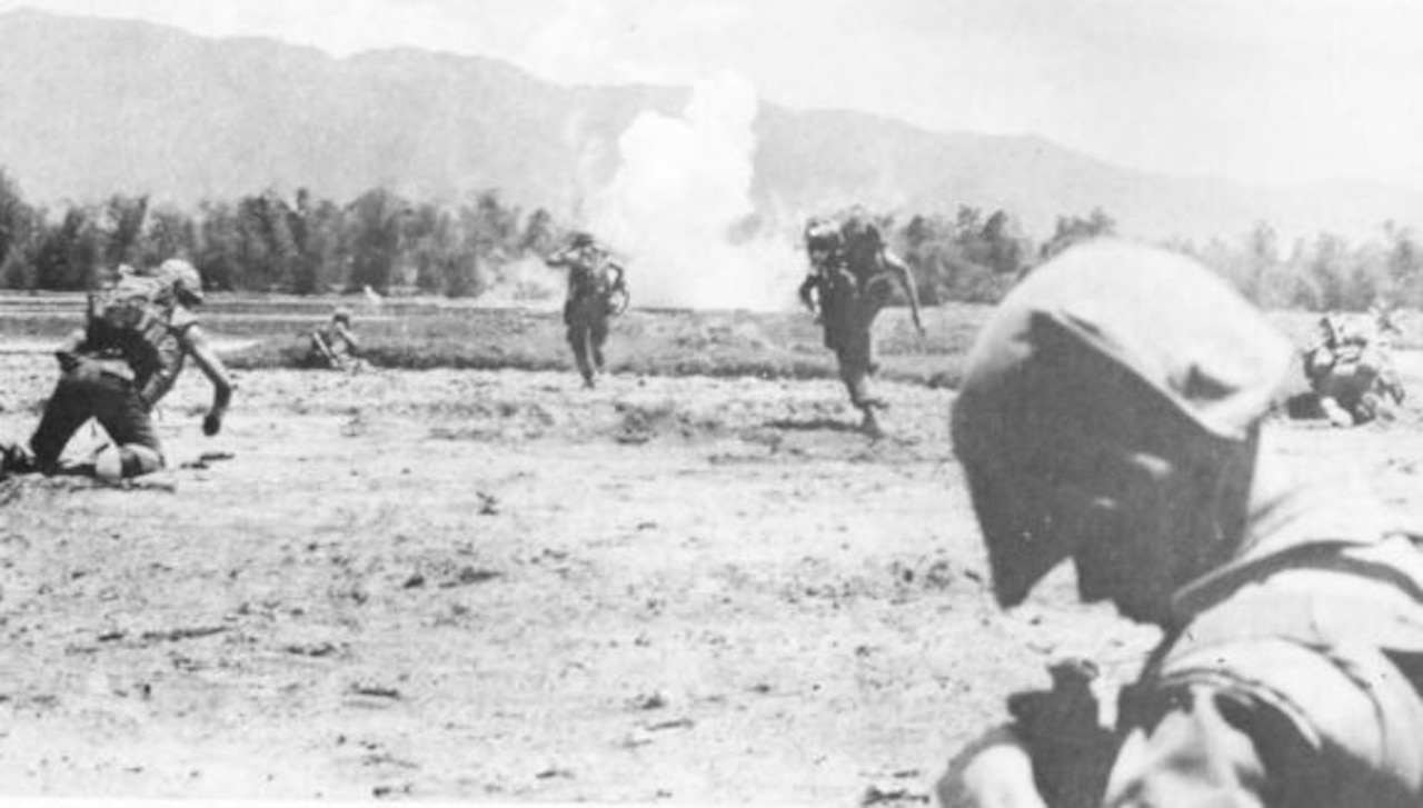 Men in combat gear run through an open field toward an explosion.