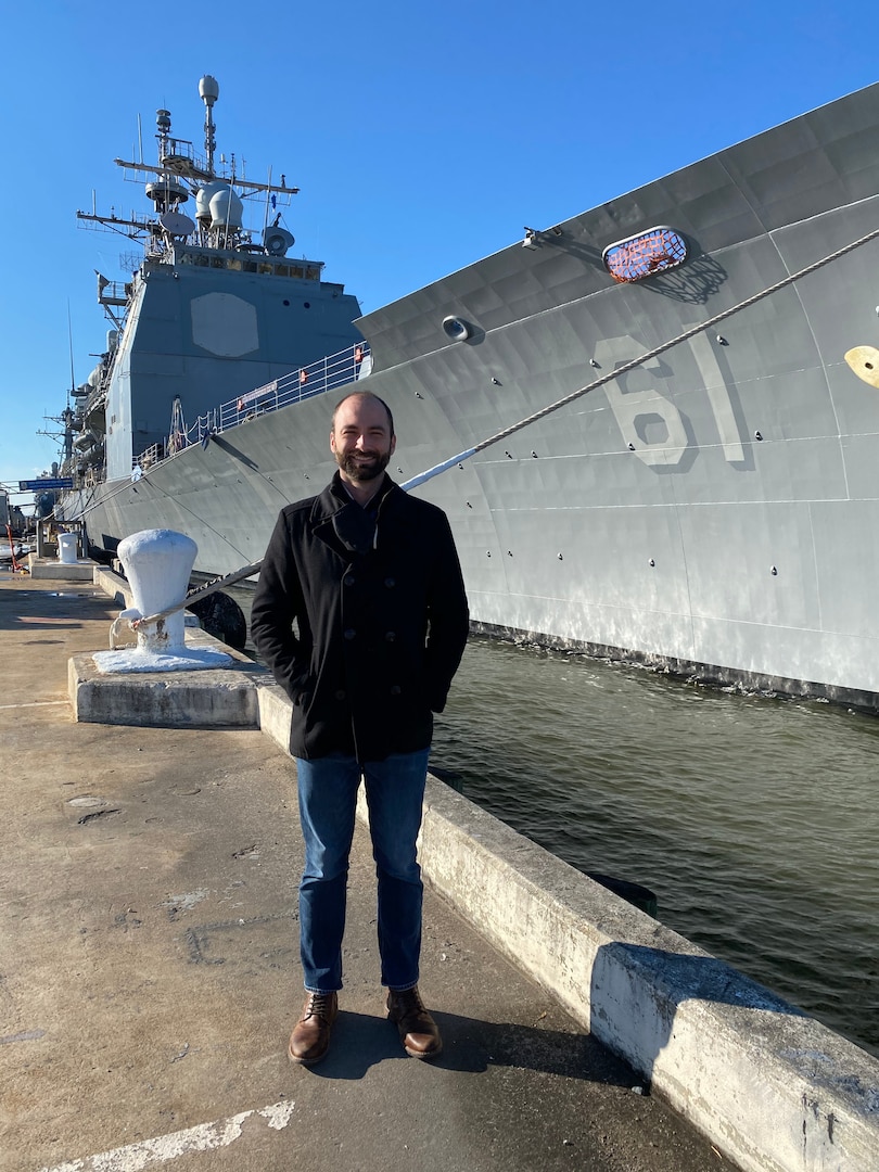 IMAGE: Naval Surface Warfare Center Dahlgren Division scientist Adam Goetz is pictured in front of the Ticonderoga-class guided-missile cruiser USS Monterey (CG 61). The new hire onboarding process was efficient for Goetz, who is a new project lead for the Human Systems Integration’s Space Design and Analysis Group. Within a matter of days after completing new hire training and orientation, Goetz utilized his experience in human factors design analysis to assist his team in development of a Bridge model for the U.S. Coast Guard at the department’s Human Performance Lab.  (U.S. Navy photo/Released)