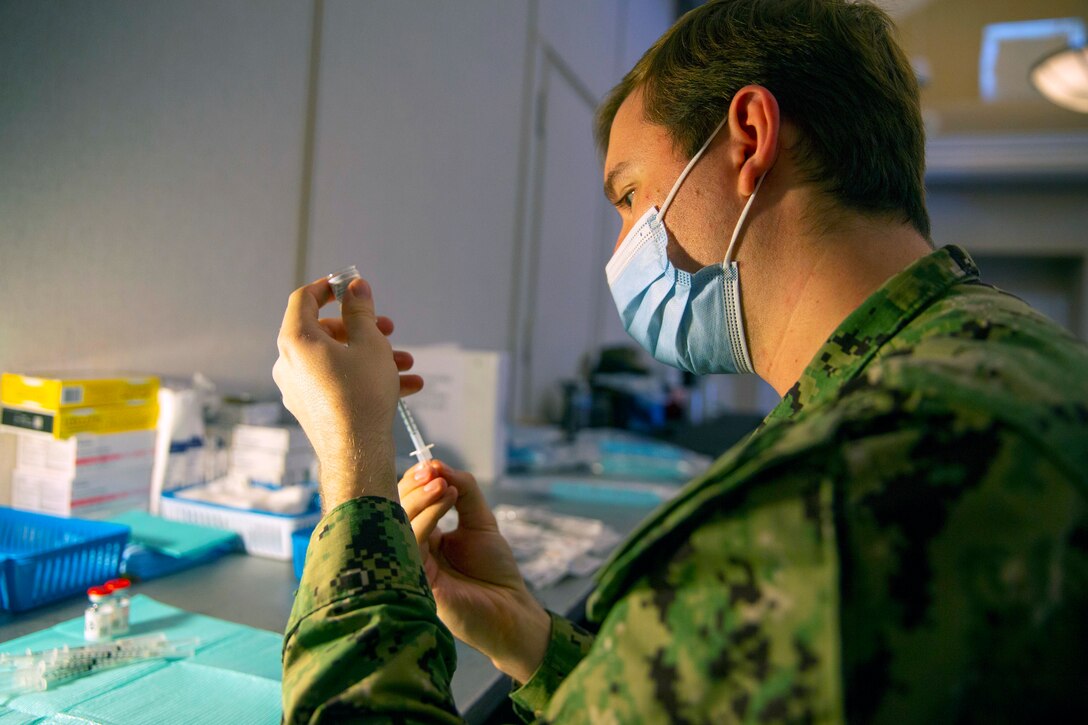 A service member wearing a face mask uses a syringe to draw fluid from a small bottle.