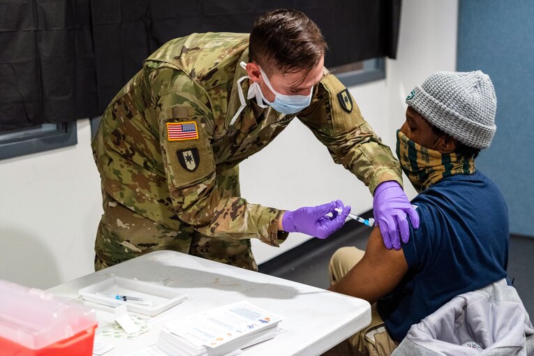 A soldier wearing a face mask and gloves leans down to give an injection to a man who's wearing a face mask seated in a chair.