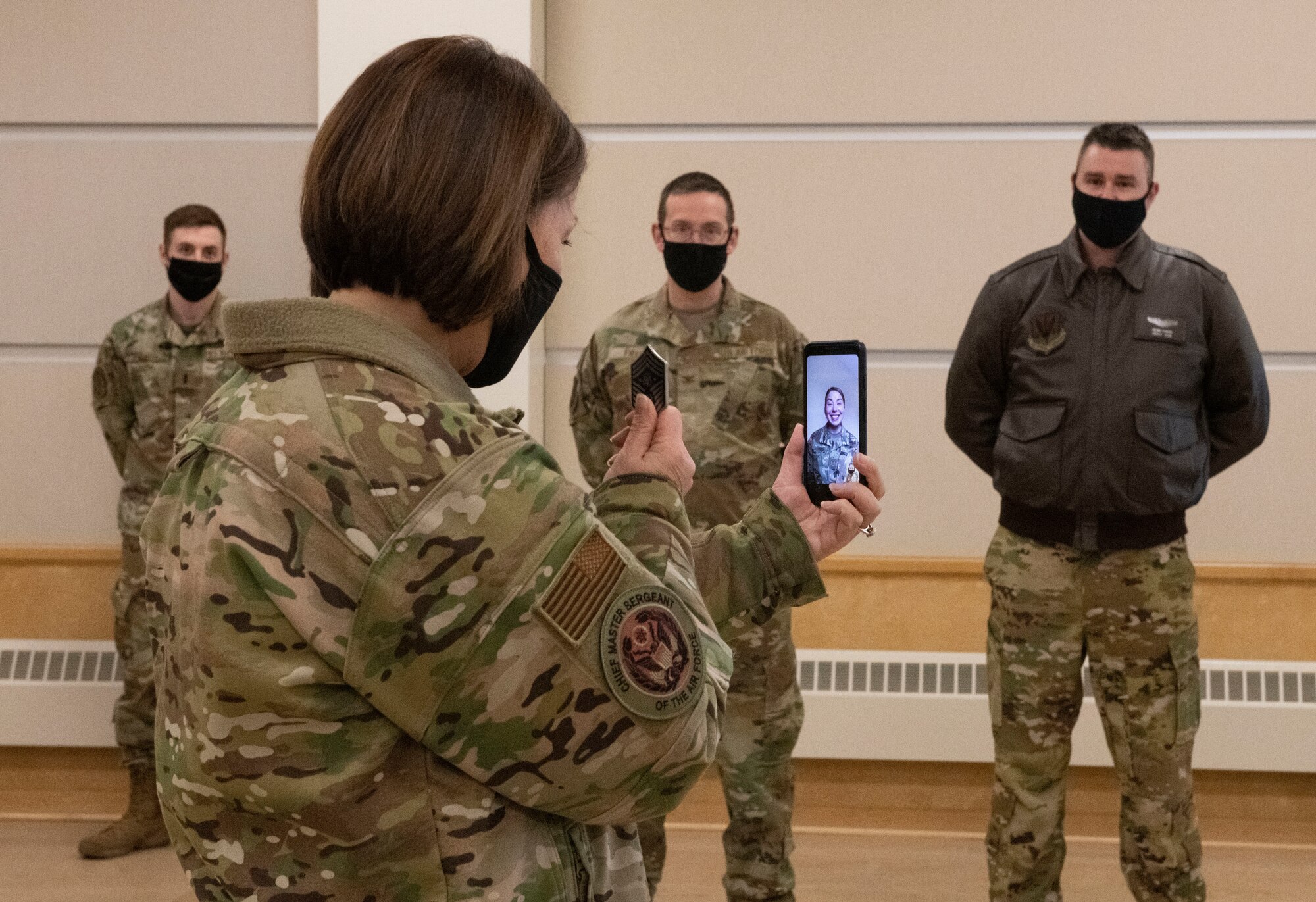 Chief Master Sergeant of the Air Force JoAnne S. Bass holds up a coin with her right hand while holding a phone with her left. On the phone is Senior Airman Gabrielle Lepinski, being virtually coined through video chat.