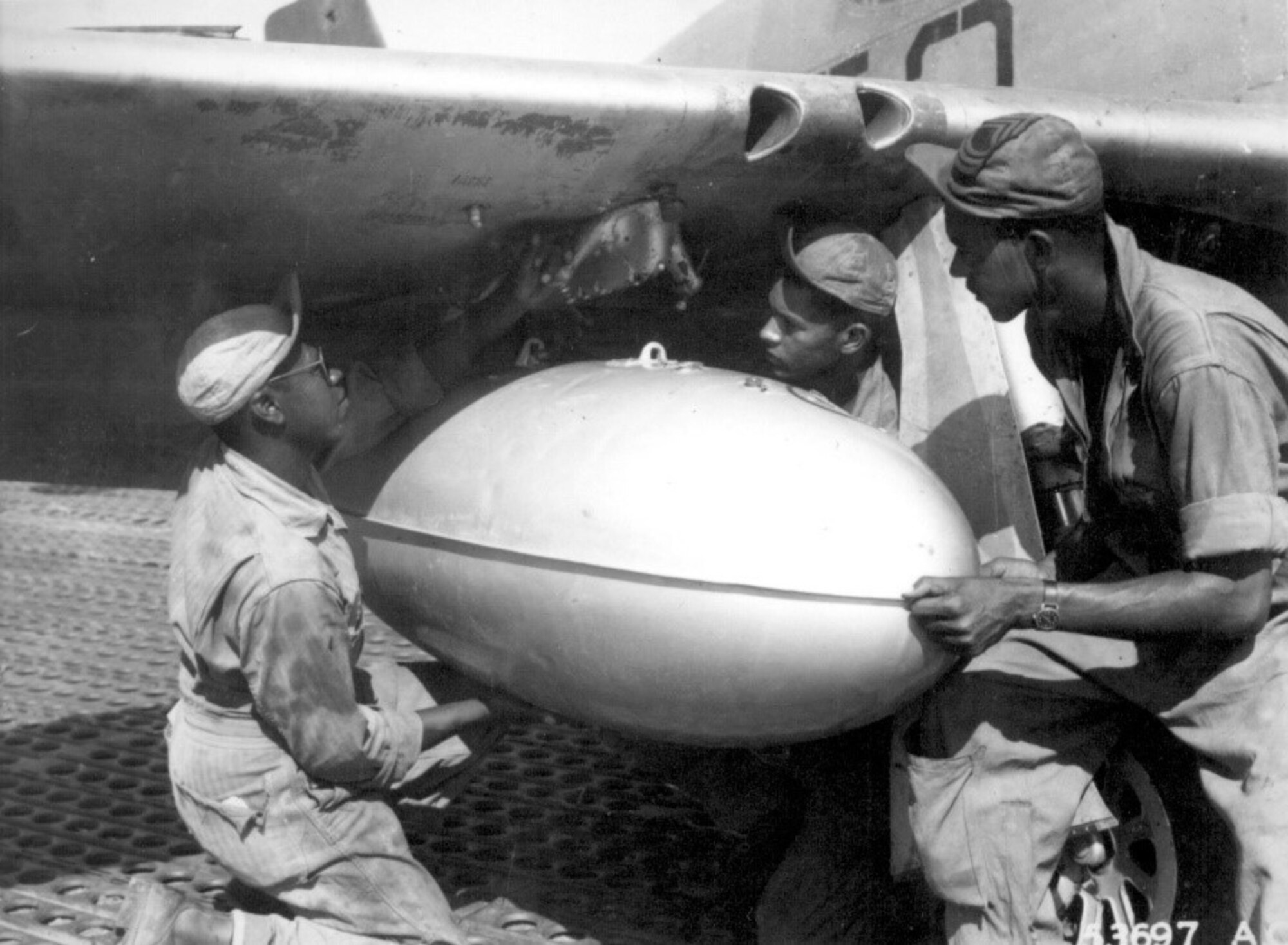 Editor’s note: photo and caption were pulled from the National Archives Catalog.
Members of the ground crew of a Negro fighter squadron of the 15th U.S. Air Force in Italy place a loaded wing tank on a P-51 Mustang before the group takes off on another mission escorting bombers over enemy targets. The squadron uses auxiliary fuel tanks for long-distance flights. Left to right: T/Sgt. Charles K. Haynes, S/Sgt. James A. Sheppard, and M/Sgt. Frank Bradley. N.d. 208-AA-49E-1-3.