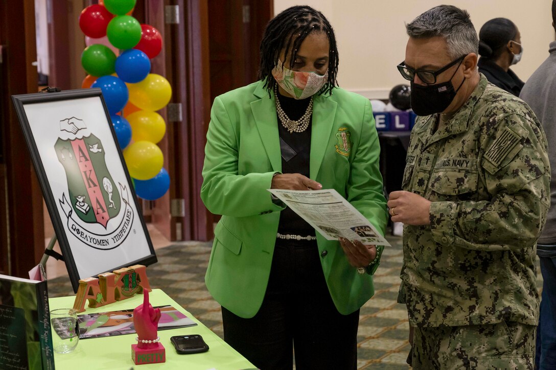 A civilian speaks with a sailor by a table at an indoor event.