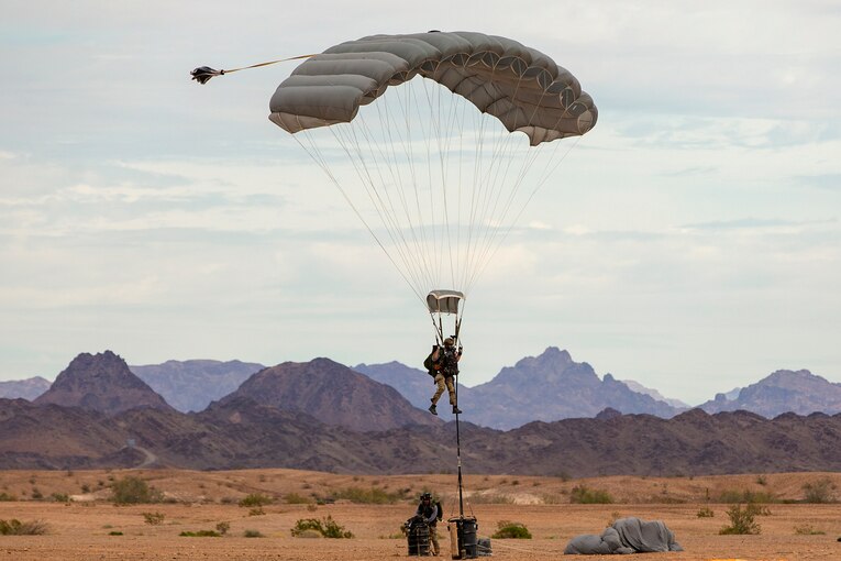 Airmen and soldiers freefall with parachutes while carrying barrels.